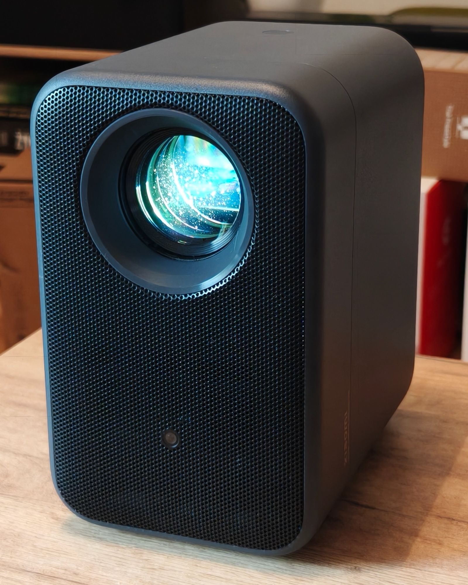 Włączony projektor Xiaomi L1 Pro ujęty pod kątem ze świecącym obiektywem i ciemną perforowaną obudową