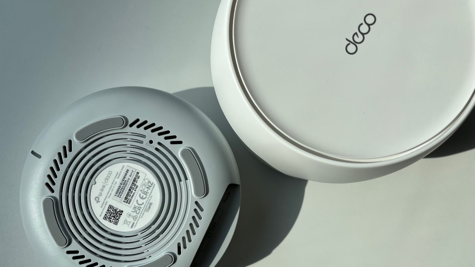 Spód routera TP-Link Deco z widocznymi otworami wentylacyjnymi i tabliczką znamionową oraz drugie urządzenie leżące obok